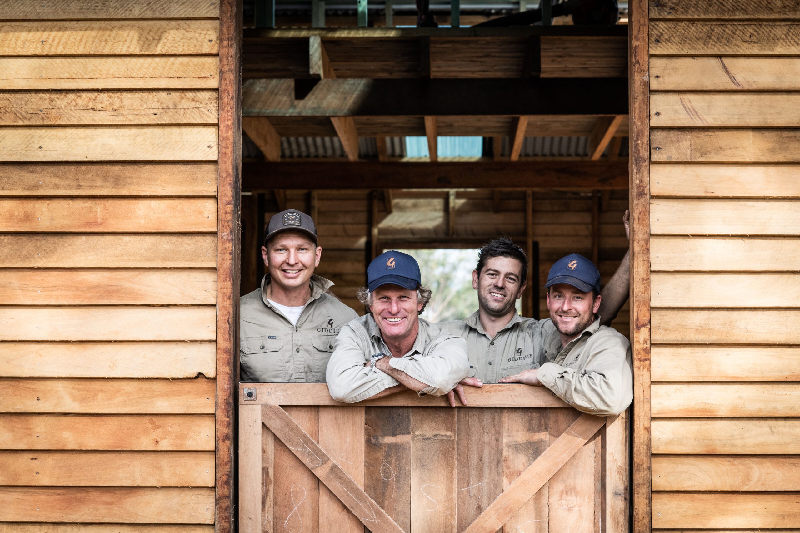 DSC_8479_1_optimized_2000 Four Giddiup team members smiling and leaning against a wooden gate, showcasing their craftsmanship in a timber structure, emphasizing expertise in post and rail fencing.