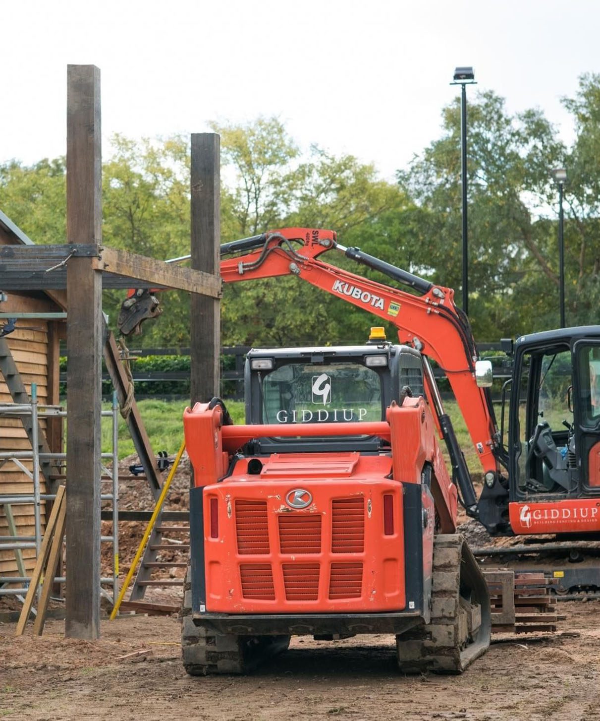 img-4 Construction site featuring Giddiup-branded machinery, including a Kubota excavator and a loader, working on timber post and rail fencing installation amidst a natural setting.